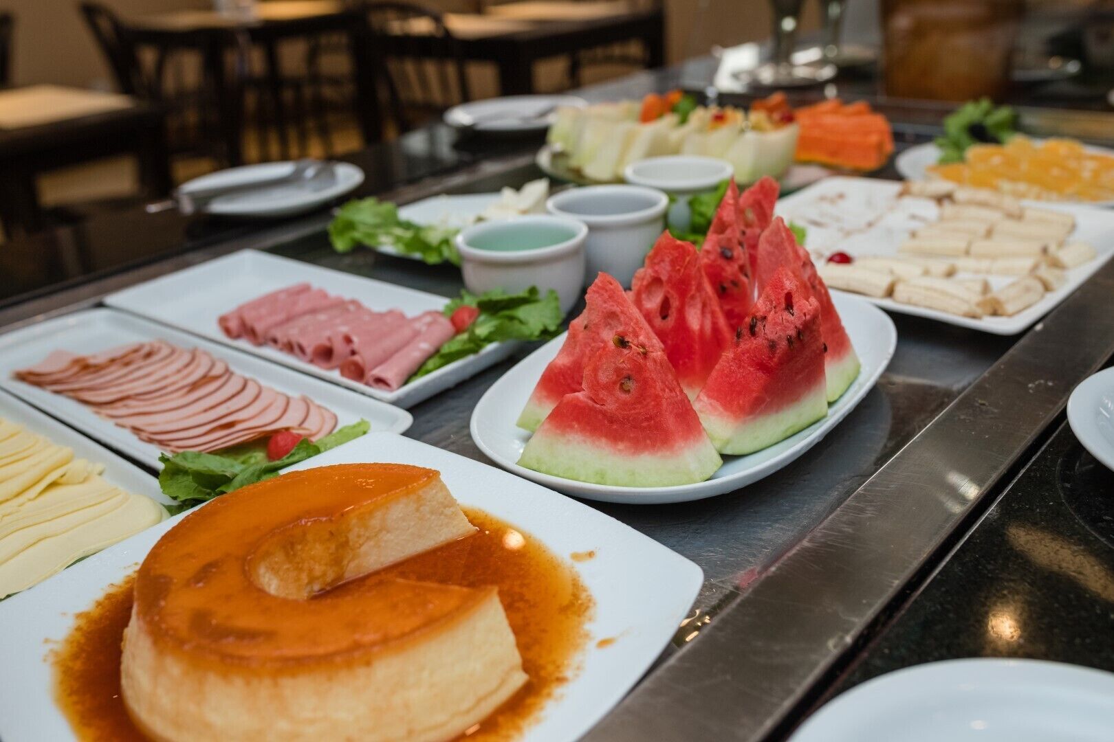 A buffet table with various food items including sliced meats, watermelon, and a flan dessert.