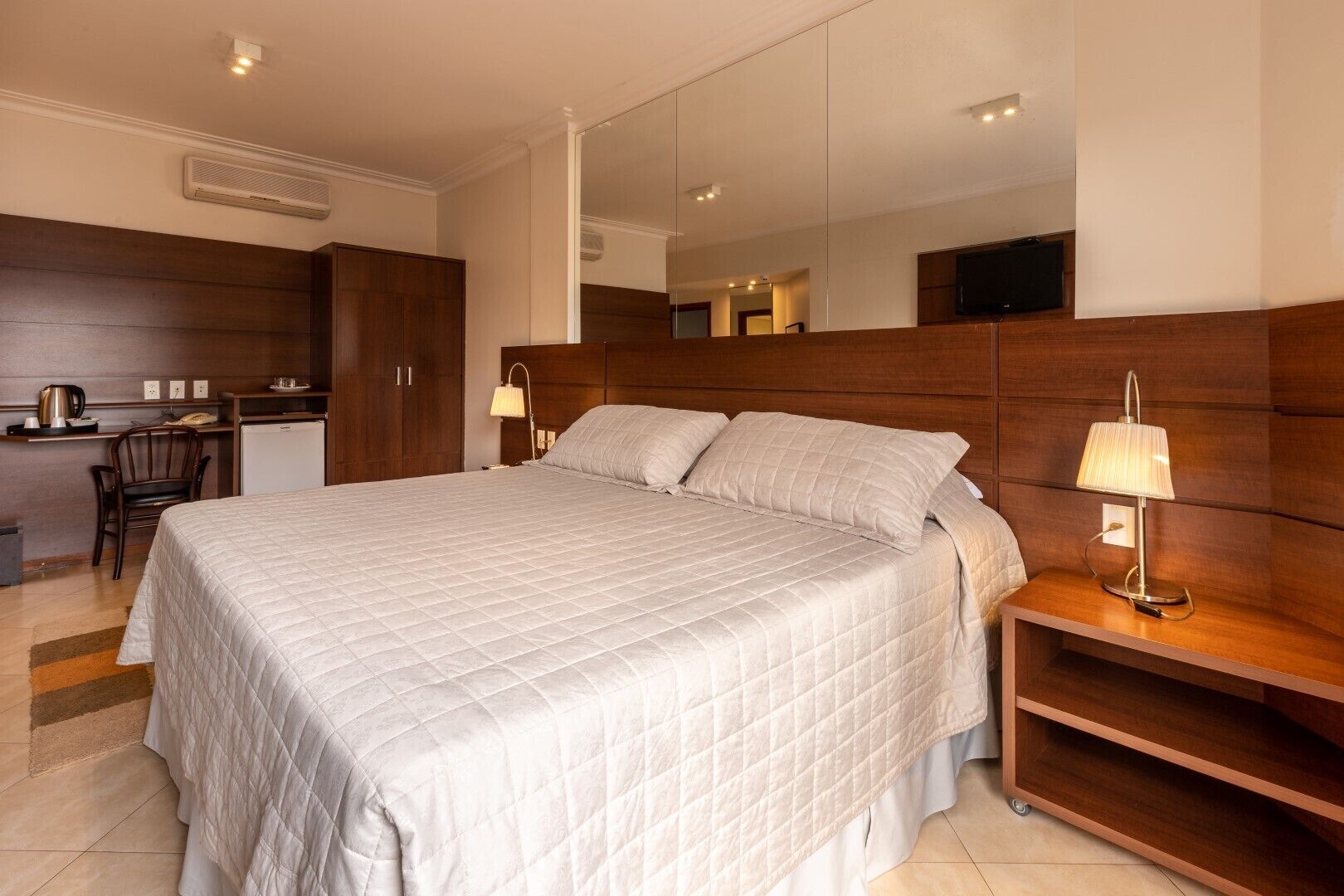 Quarto de hotel com uma cama grande, uma escrivaninha com cadeira, um espelho e uma televisão.