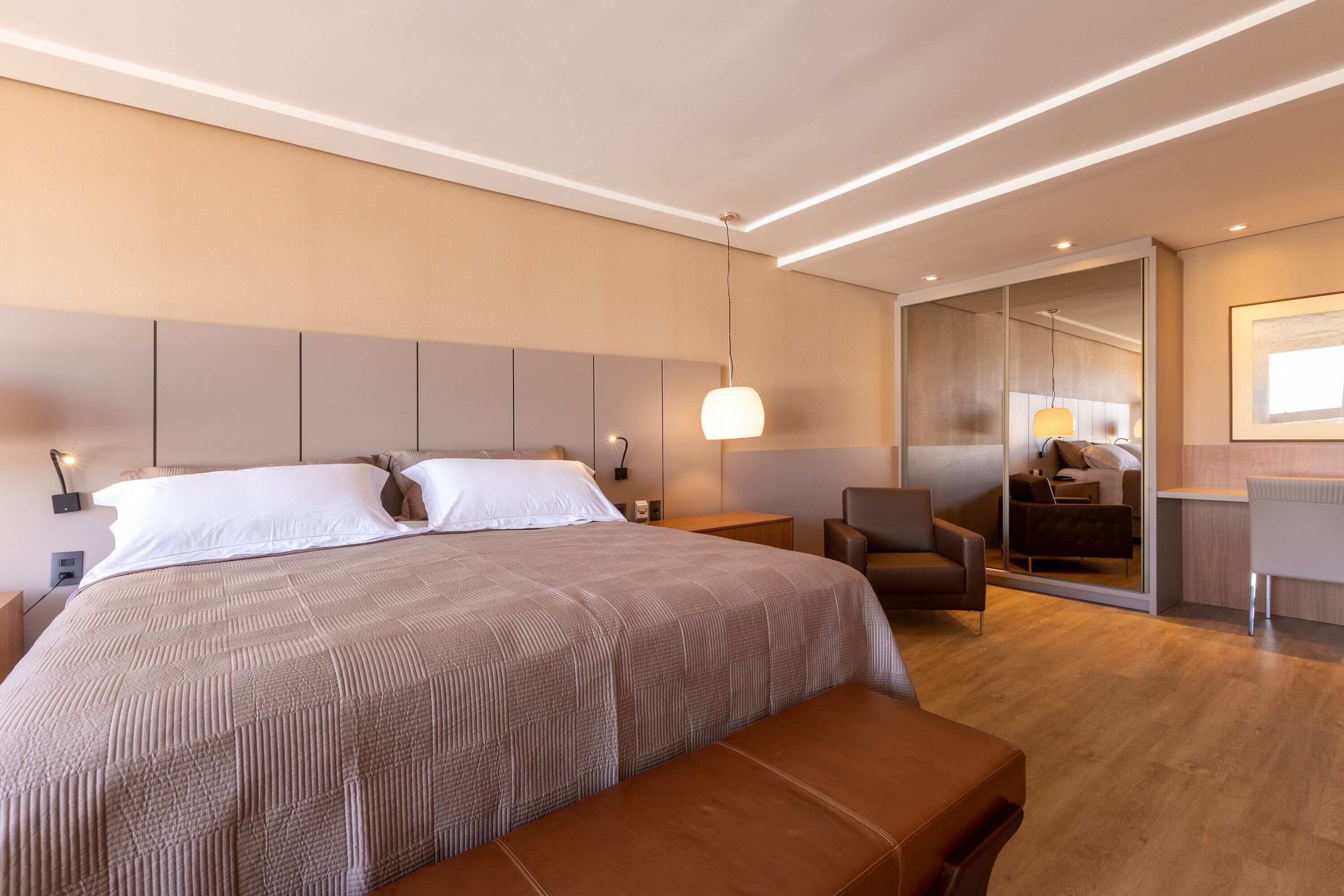 A modern hotel room with a large bed, a sitting area with a chair, a desk, and a mirror.