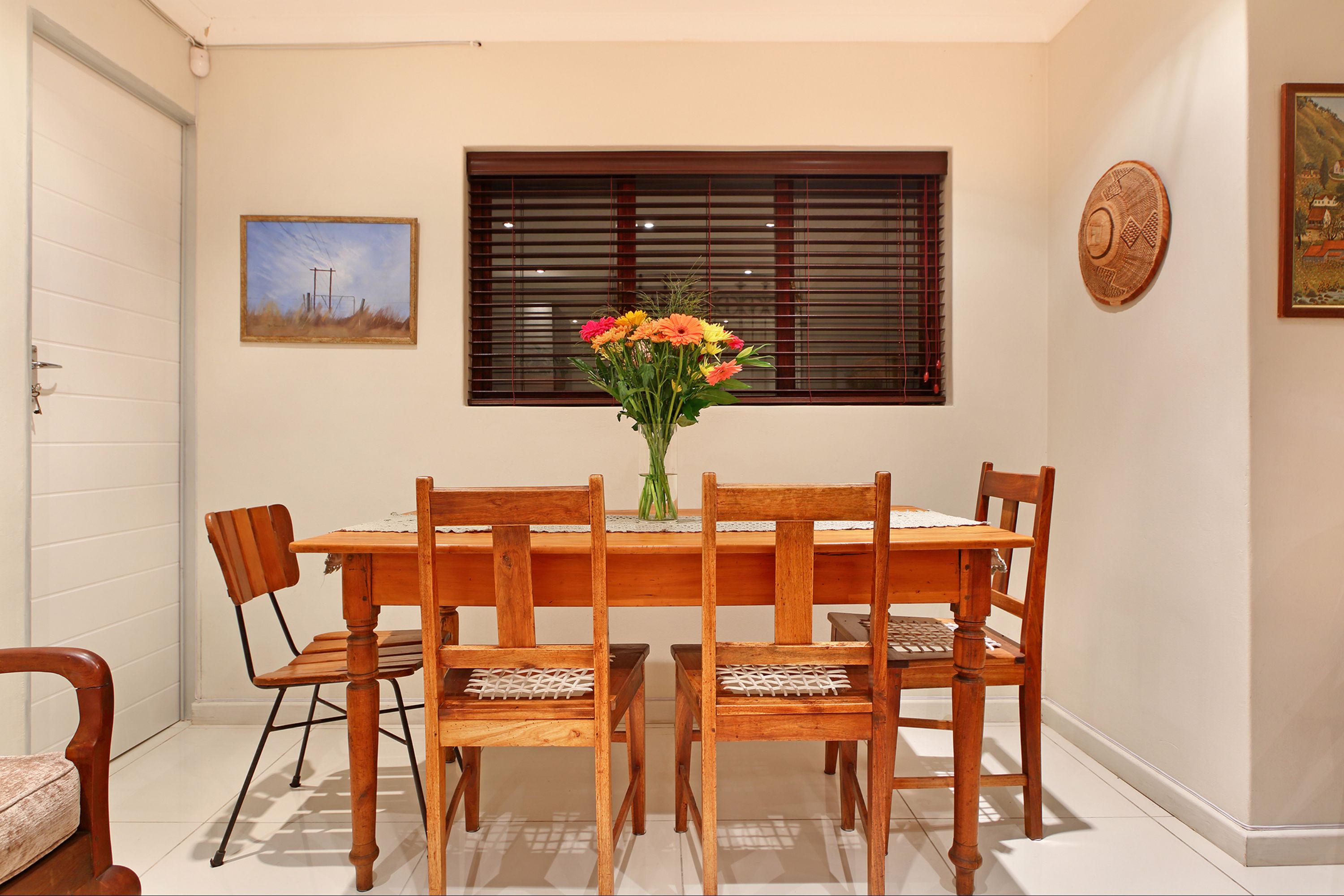 A dining area with a wooden table and chairs, a vase with flowers, and a window with blinds.