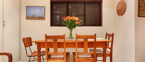 A dining area with a wooden table and chairs, a vase with flowers, and a window with blinds.