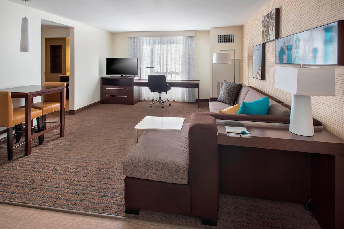A hotel room with a brown sofa, a wooden desk, a television, and a lamp.