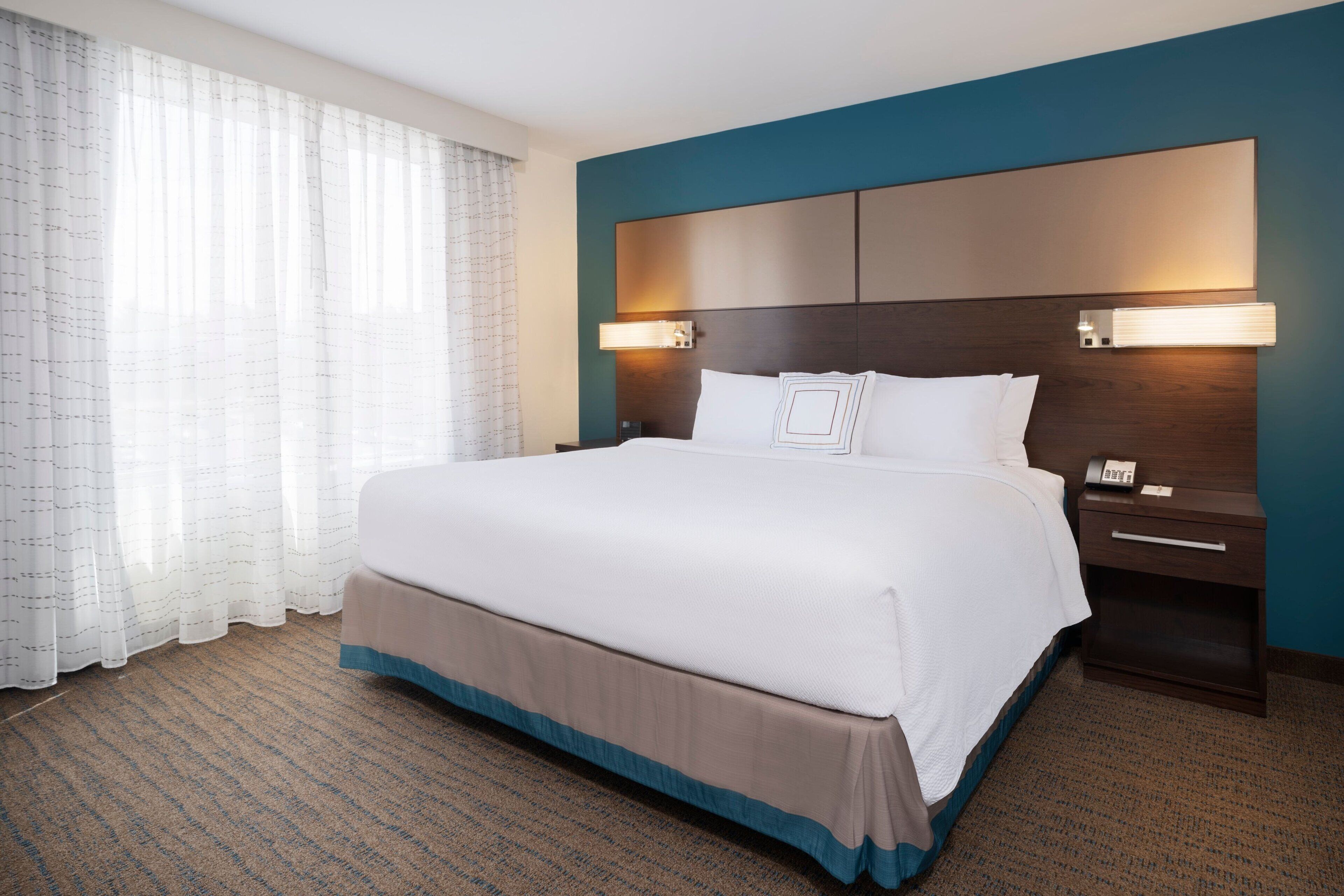 A hotel room with a large bed, a nightstand with a phone, and a headboard with wall-mounted lights.