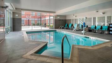 An indoor swimming pool with clear glass walls, multiple lounge chairs, and a view of a red brick building outside.