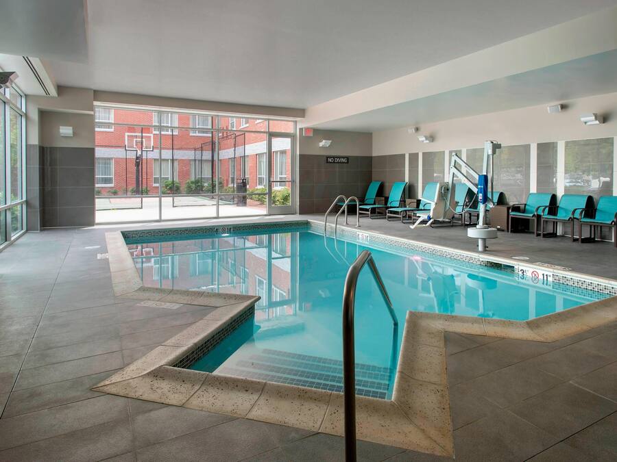 An indoor swimming pool with clear glass walls, multiple lounge chairs, and a view of a red brick building outside.