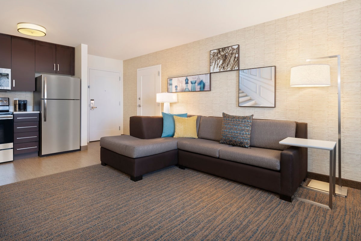 A modern living room with a sectional sofa, a flat-screen TV, and a kitchen area with a refrigerator and microwave.