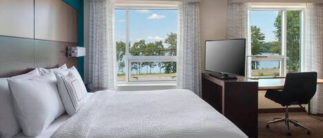 Habitación de hotel con una cama grande, un escritorio con silla, televisión y ventana con vistas a árboles y un cuerpo de agua.