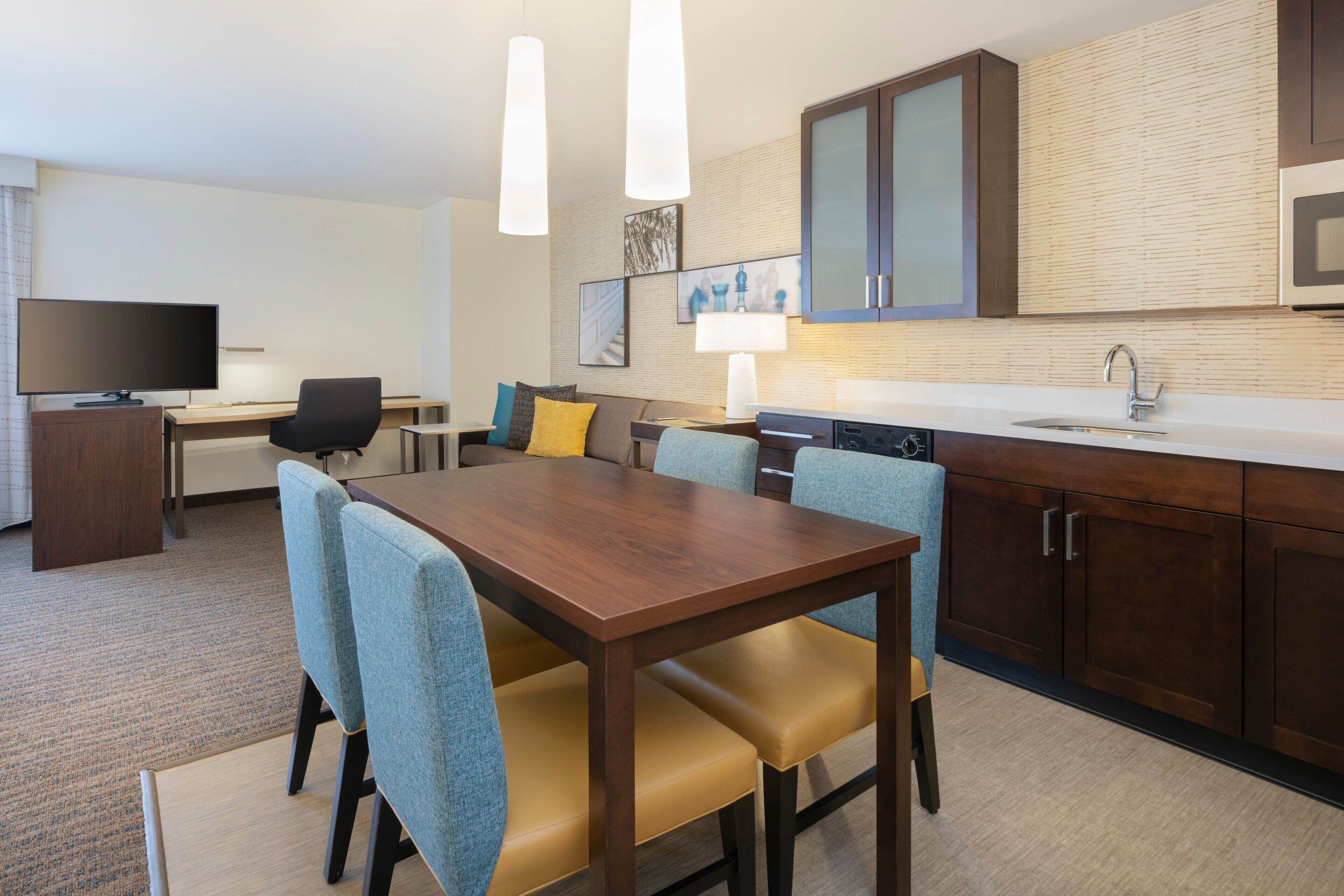 Habitación de hotel con una mesa de comedor de madera, sillas tapizadas en azul, un televisor de pantalla plana, un escritorio y una kitchenette con microondas y lavaplatos.