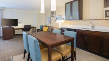 Habitación de hotel con una mesa de comedor de madera, sillas tapizadas en azul, un televisor de pantalla plana, un escritorio y una kitchenette con microondas y lavaplatos.