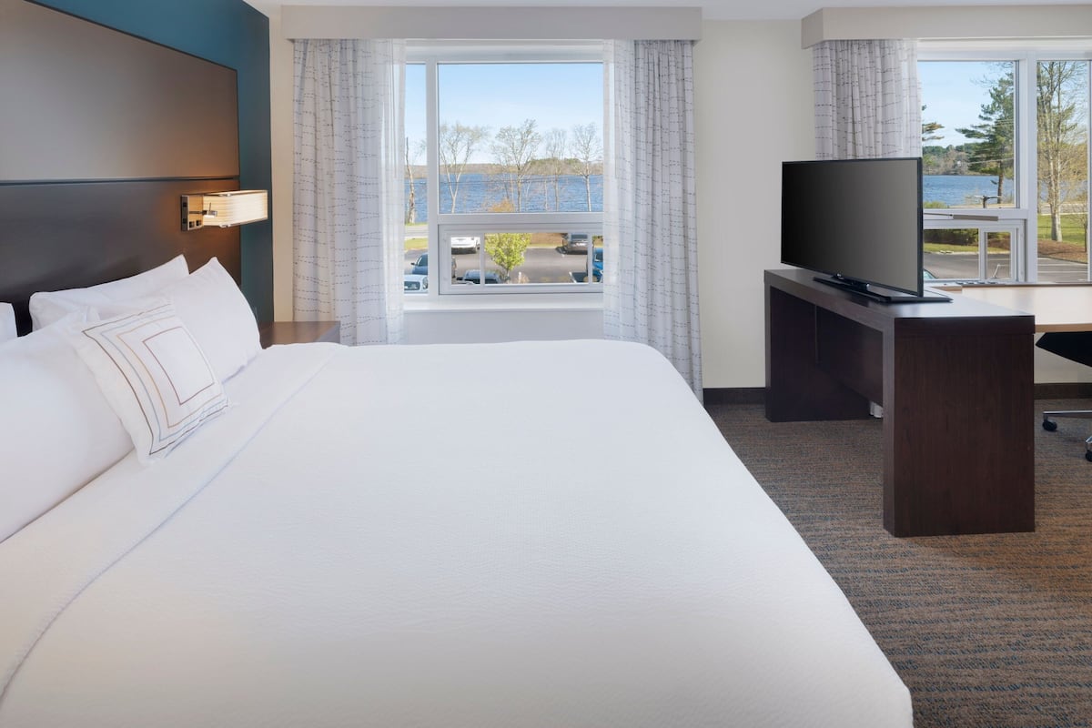 A hotel room with a large bed, a desk with a television, and a window with a view of a body of water and trees.