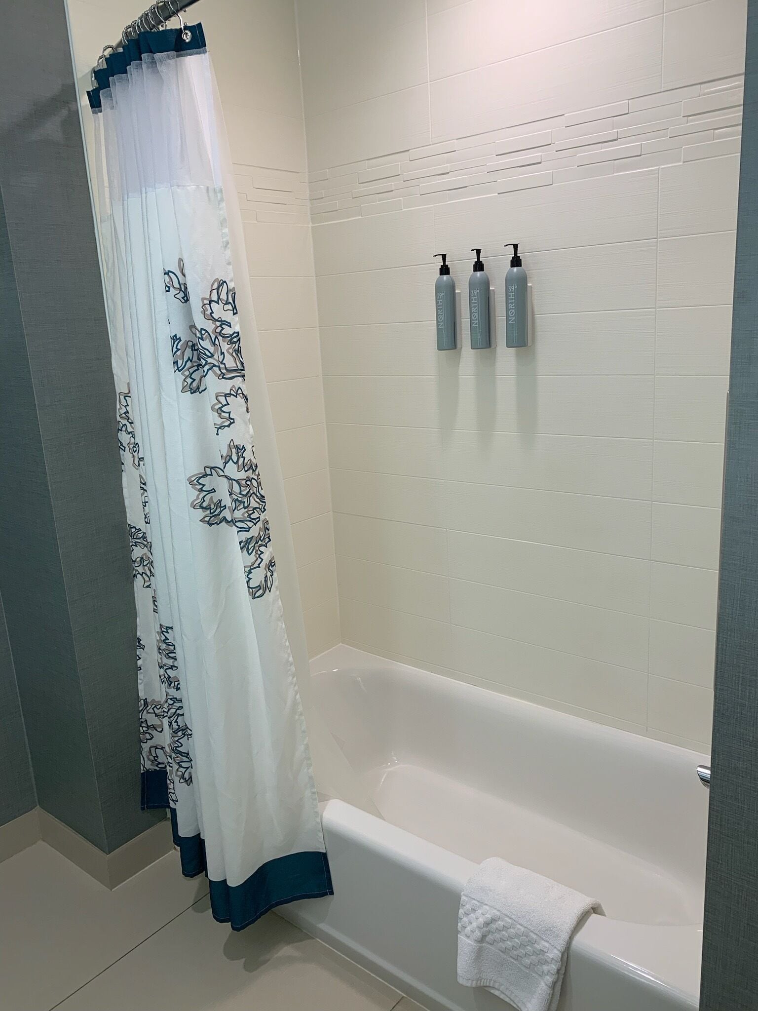 A bathroom with a bathtub, a shower curtain with a floral pattern, and three bottles of shampoo on the wall.
