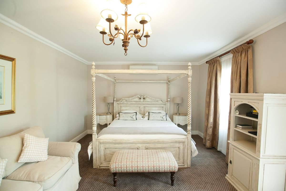 Ein Bett mit Baldachin, ein weißes Kopfteil, eine Bank am Fußende, ein Nachttisch mit Lampe und ein Schrank mit Regalen.