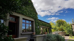 Une maison avec un toit à forte pente recouvert de lierre, de grandes fenêtres et un jardin bien entretenu agrémenté de plantes en pot.