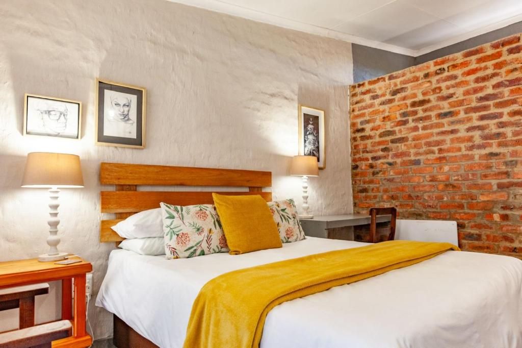 A hotel room with a brick wall, a wooden headboard, a bed with white bedding and a yellow throw, and two bedside lamps.