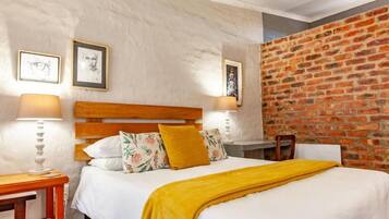 A hotel room with a brick wall, a wooden headboard, a bed with white bedding and a yellow throw, and two bedside lamps.