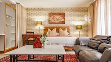 A hotel room with a brick-patterned wall, a bed with a wooden headboard, a sofa, a table with a vase, and a lamp.