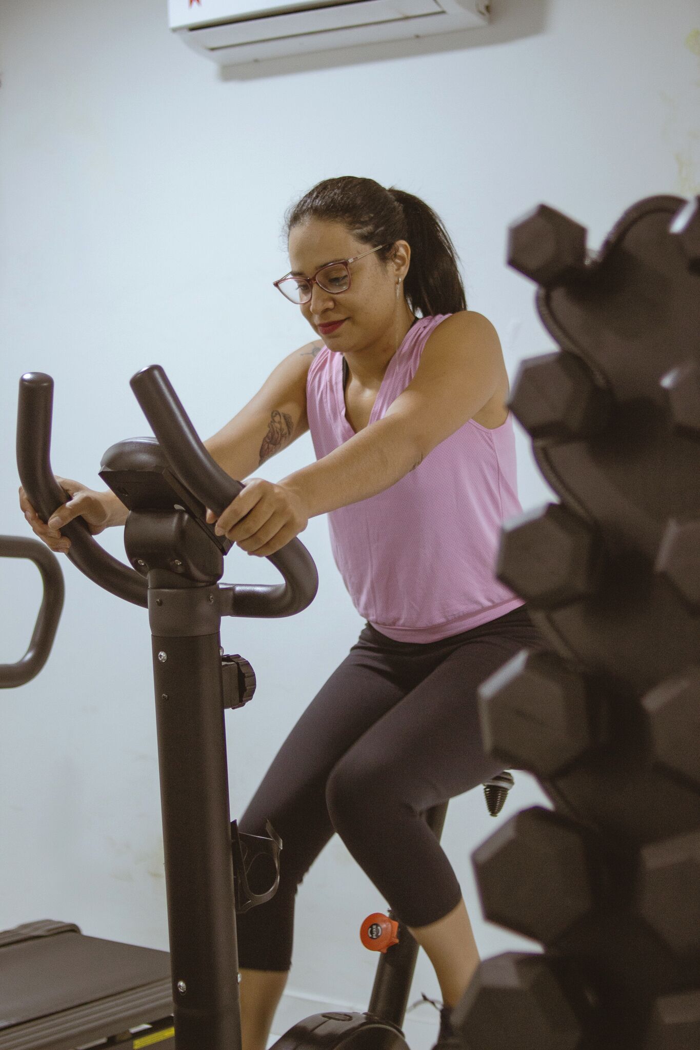 A person using an exercise bike in a room with a white wall and a large set of dumbbells in the background.