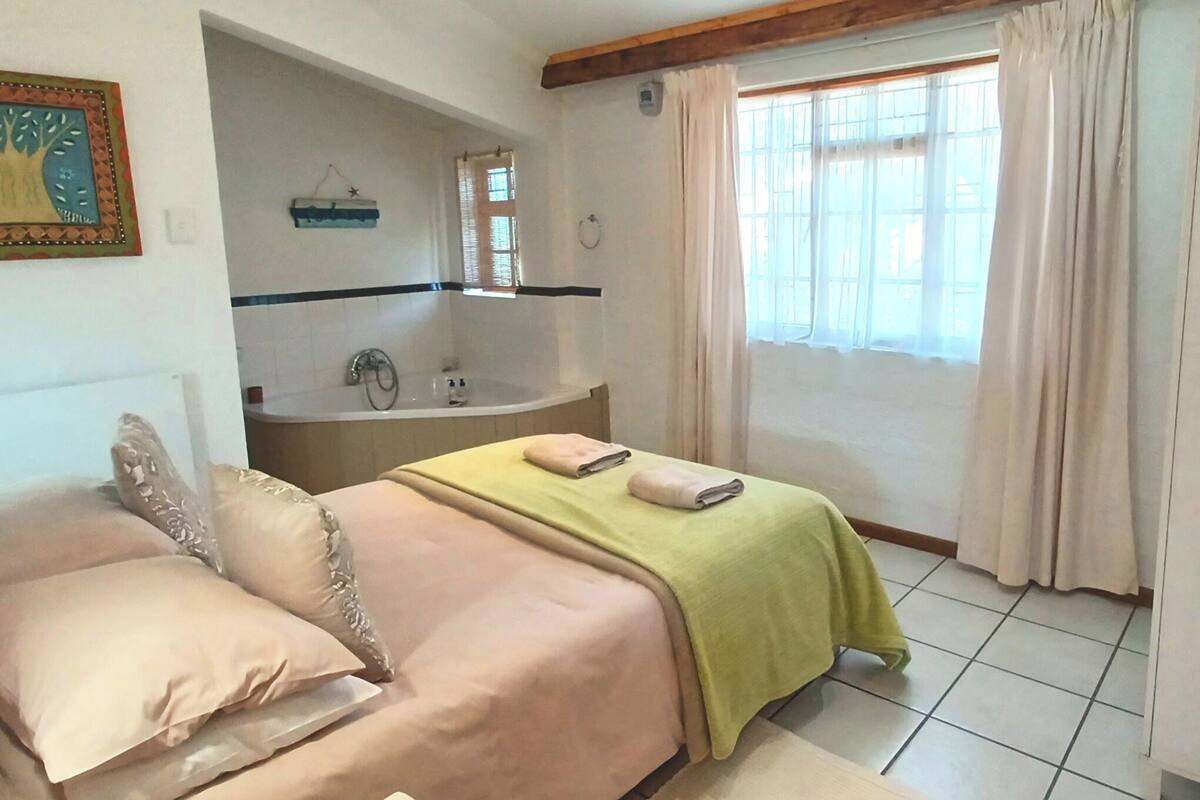 A bedroom with a bed, a bedside table, a window with curtains, a ceiling fan, and a bathroom with a bathtub.