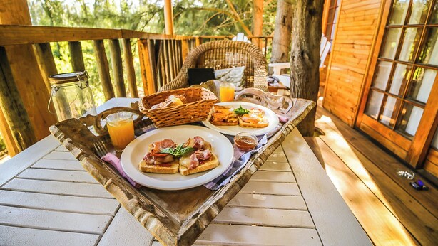 Une terrasse en bois avec une table mise pour le petit-déjeuner, comprenant des toasts, un panier et un verre de jus d’orange.