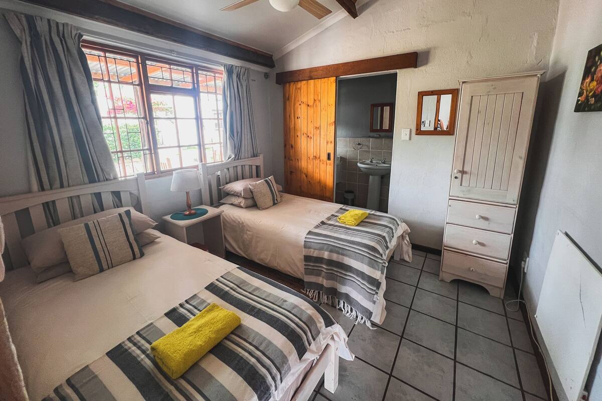 A room with two beds, a ceiling fan, a window with curtains, a sink, and a cabinet.