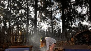 Entorno rústico al aire libre con chimenea de piedra, sillones de madera y una persona cuidando el fuego.