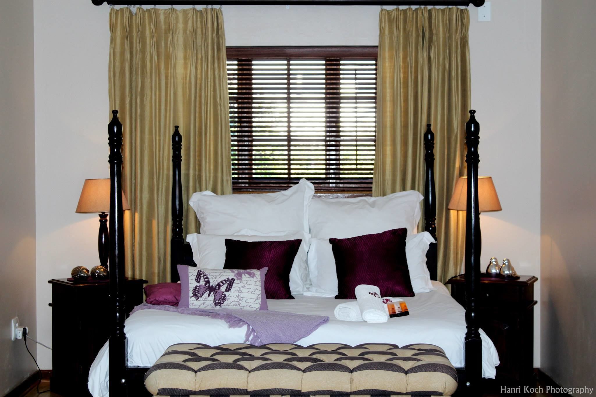 Una cama con dosel en tonos blanco y morado, dos mesitas de noche con lámparas, y una ventana con persianas.