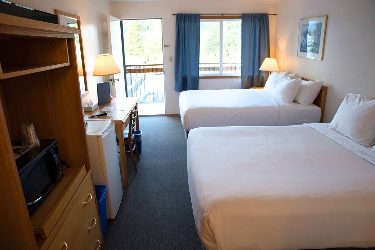 Habitación de hotel con dos camas, un escritorio con lámpara, microondas y vistas al exterior a través de una puerta corrediza.