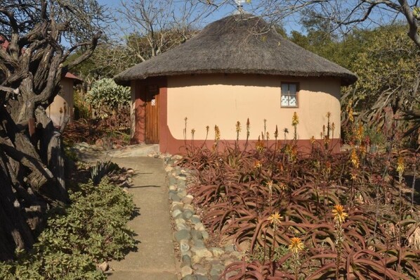 A thatched-roof hut with a small window, surrounded by a garden with various plants and a stone pathway. - uMuzi Ondini (Ulundi)