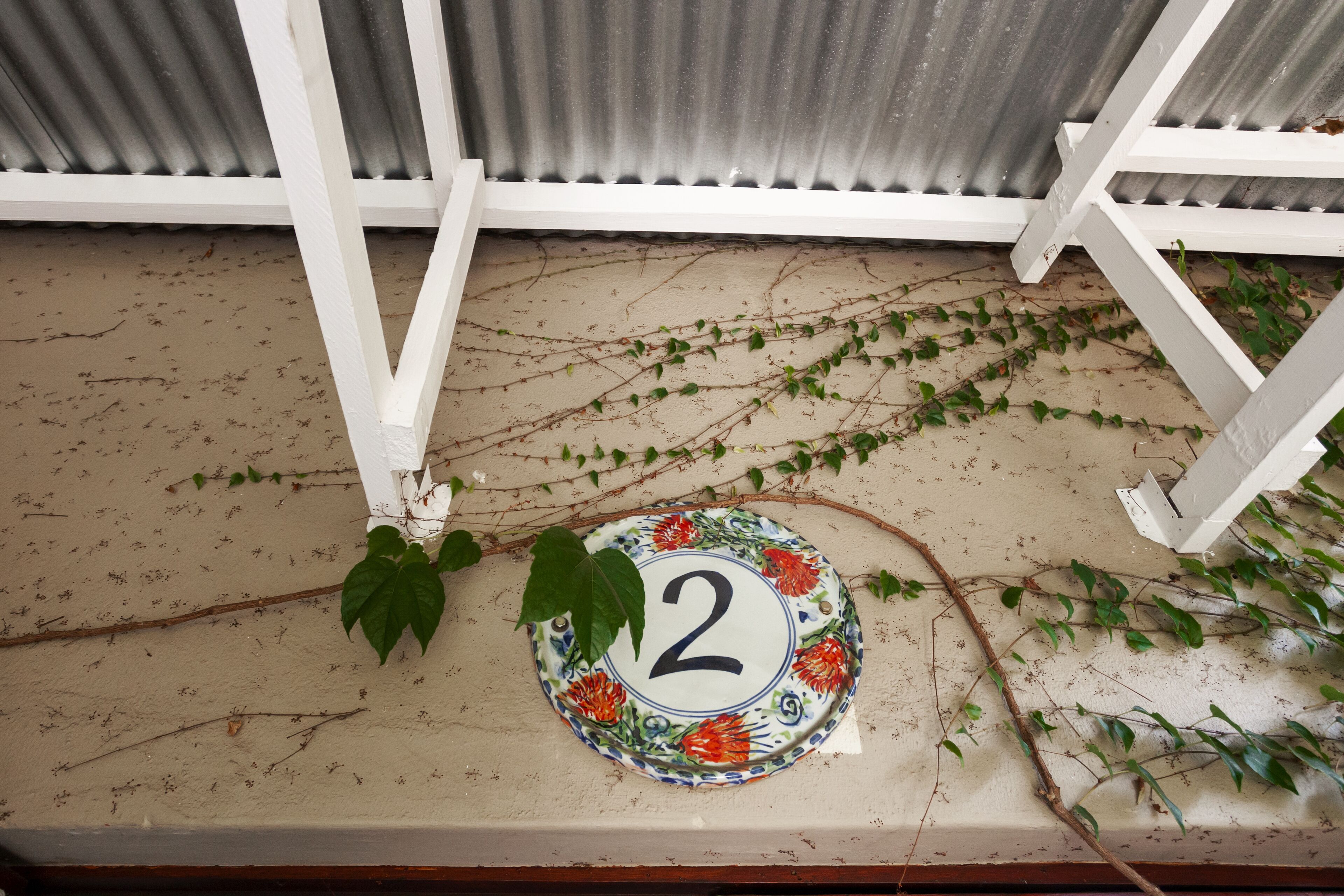 A house number '2' on a decorative plate, surrounded by vines, with a corrugated metal wall and white wooden beams in the background.