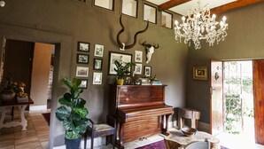 Huoneessa on puinen piano, kattokruunu, seinällä olevia kehystettyjä tauluja ja suuri huonekasvi.
