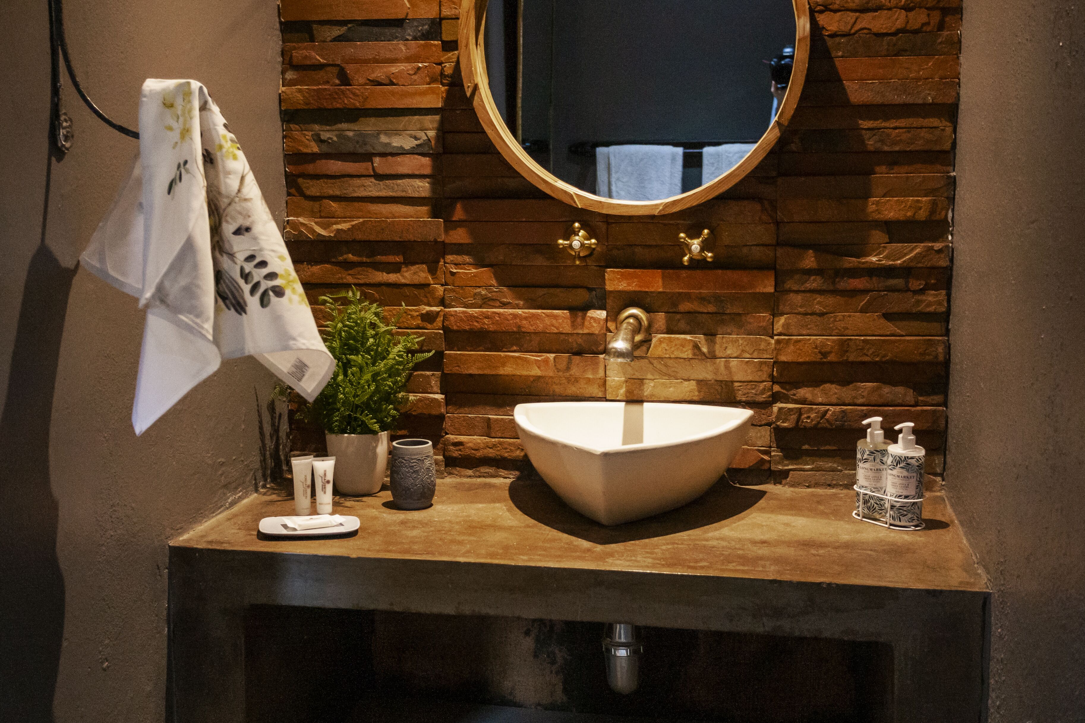 Een badkamer met een stenen muur, een ronde spiegel, een witte wastafel, een handdoek, een potplant en toiletartikelen.