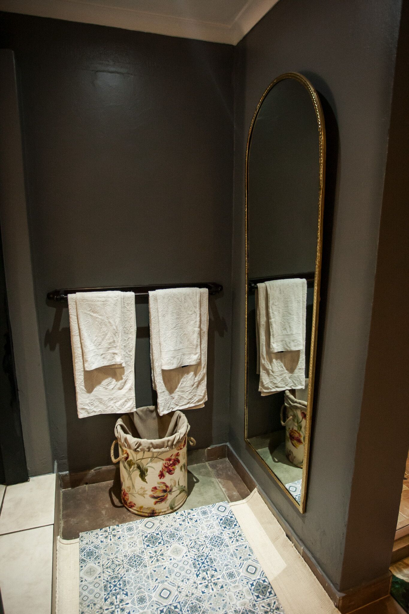 A bathroom with dark walls, a large mirror, two towels hanging on a towel rack, and a floral-patterned bucket on the floor.