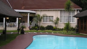 A swimming pool with a curved edge, surrounded by a brick pathway. There are two thatched-roof structures, one on each side of the pool. - Victorian Guest House (Krugersdorp)