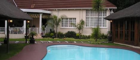 A swimming pool with a curved edge, surrounded by a brick pathway. There are two thatched-roof structures, one on each side of the pool.