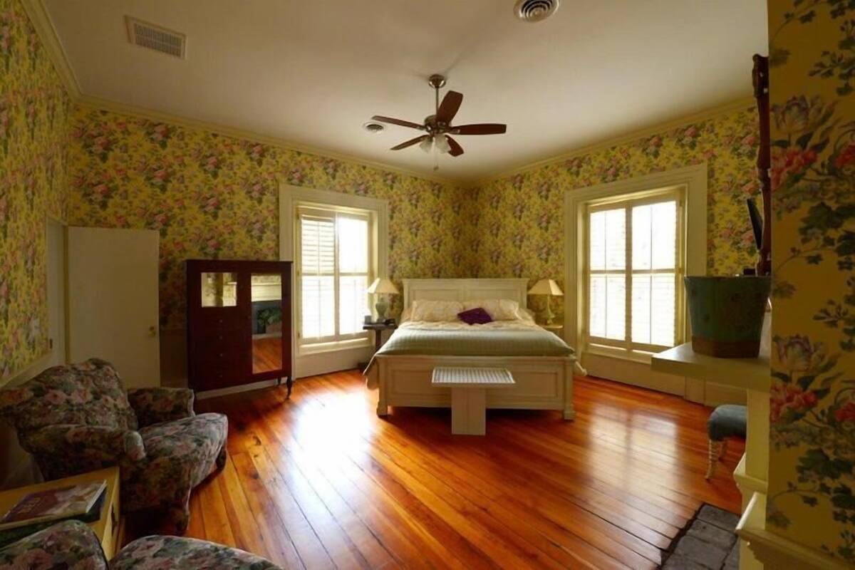Un dormitorio con piso de madera, papel tapiz floral, una cama con cabecera, dos ventanas con contraventanas, un ventilador de techo y una mesita de noche con lámpara.