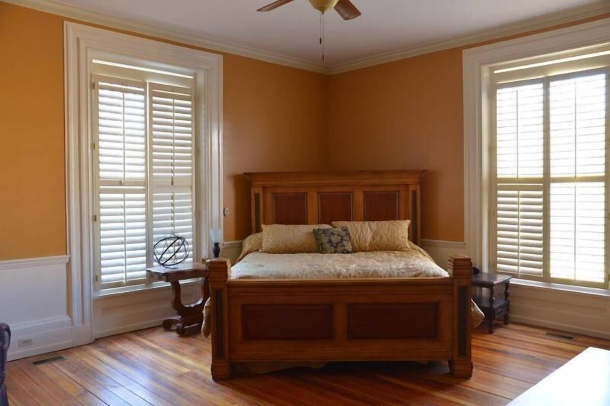 Un dormitorio con una cama de madera, dos ventanas con contraventanas y un ventilador de techo.