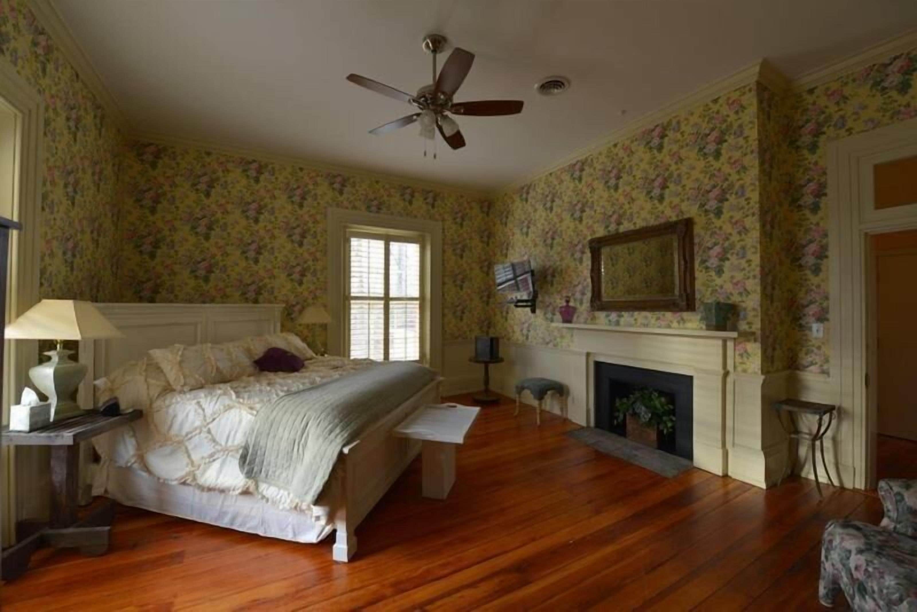 A bedroom with a bed, a ceiling fan, a fireplace, and floral wallpaper.