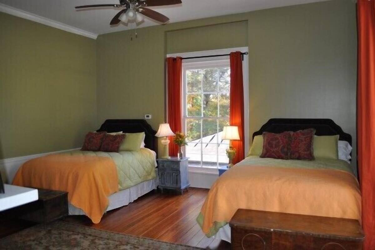 Una habitación con dos camas, un ventilador de techo, una ventana con cortinas rojas y vistas a árboles.