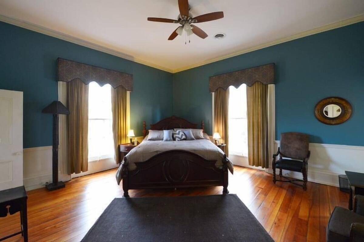 Un dormitorio con una cama de madera, dos mesitas de noche, una silla, un ventilador de techo y un amplio ventanal con cortinas.