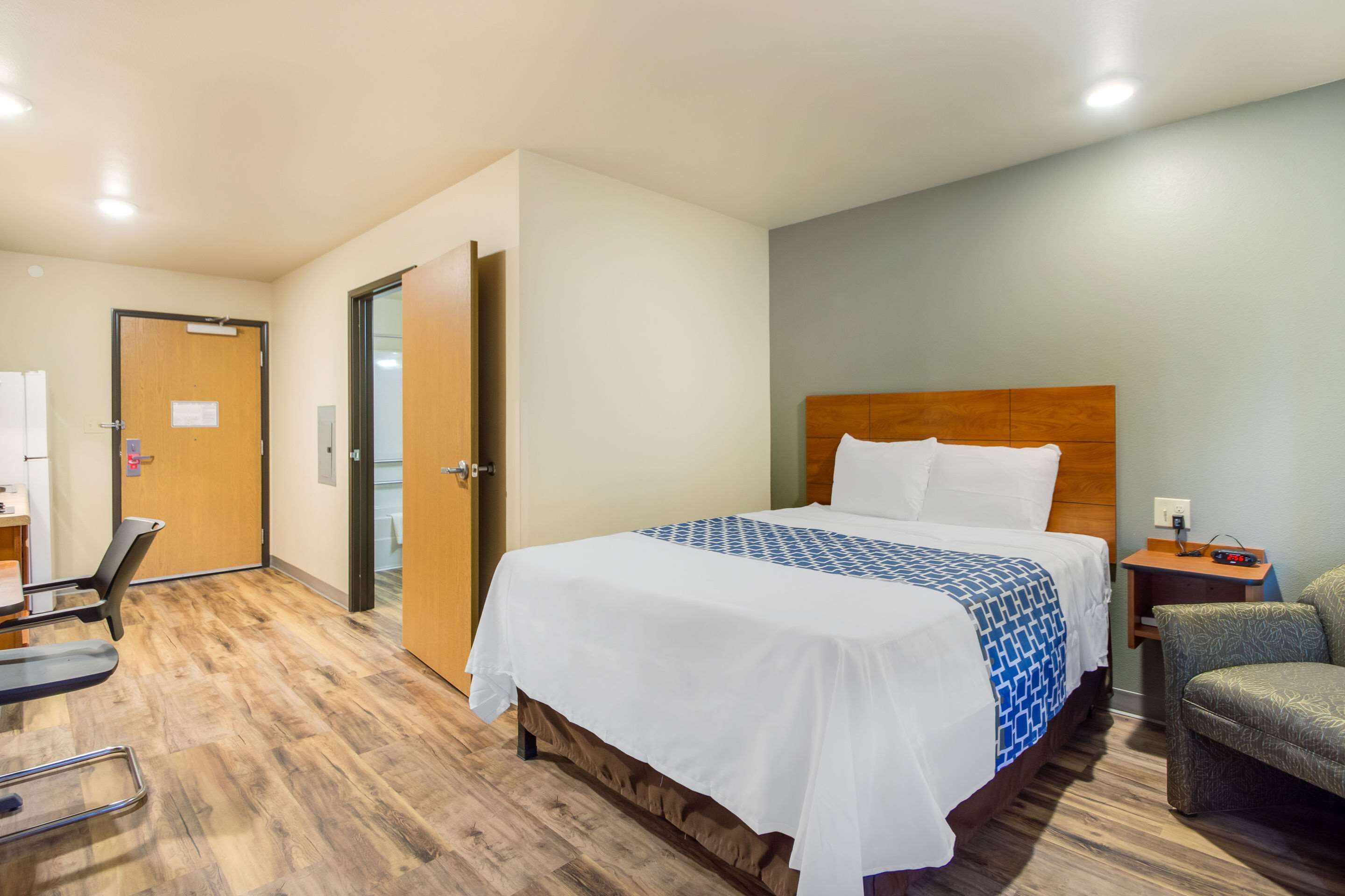A hotel room with a large bed, a wooden headboard, a desk with a chair, and a small bedside table with a lamp.