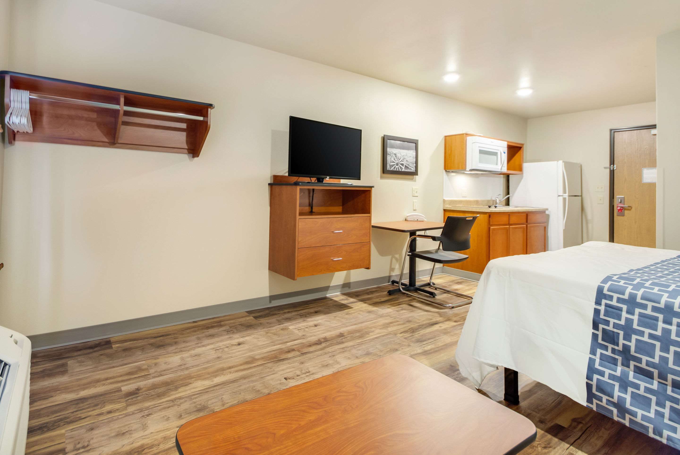 A hotel room with a bed, a desk with a chair, a television, a microwave, a refrigerator, and a wooden shelf.