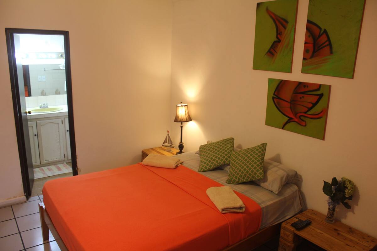 Un dormitorio con cama, almohadas, mesita de noche, lámpara y cuadros en la pared.