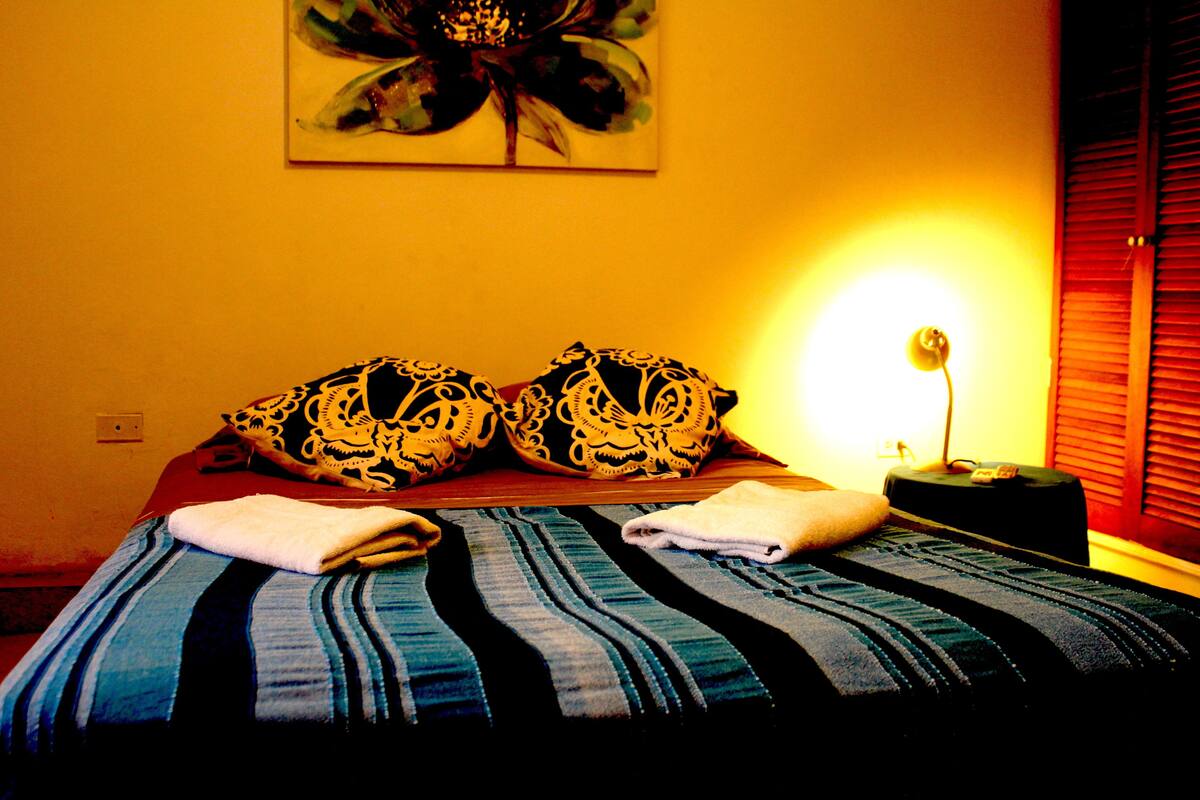 Una cama con una colcha a rayas, dos almohadas decorativas, una lámpara de noche y una obra de arte en la pared.