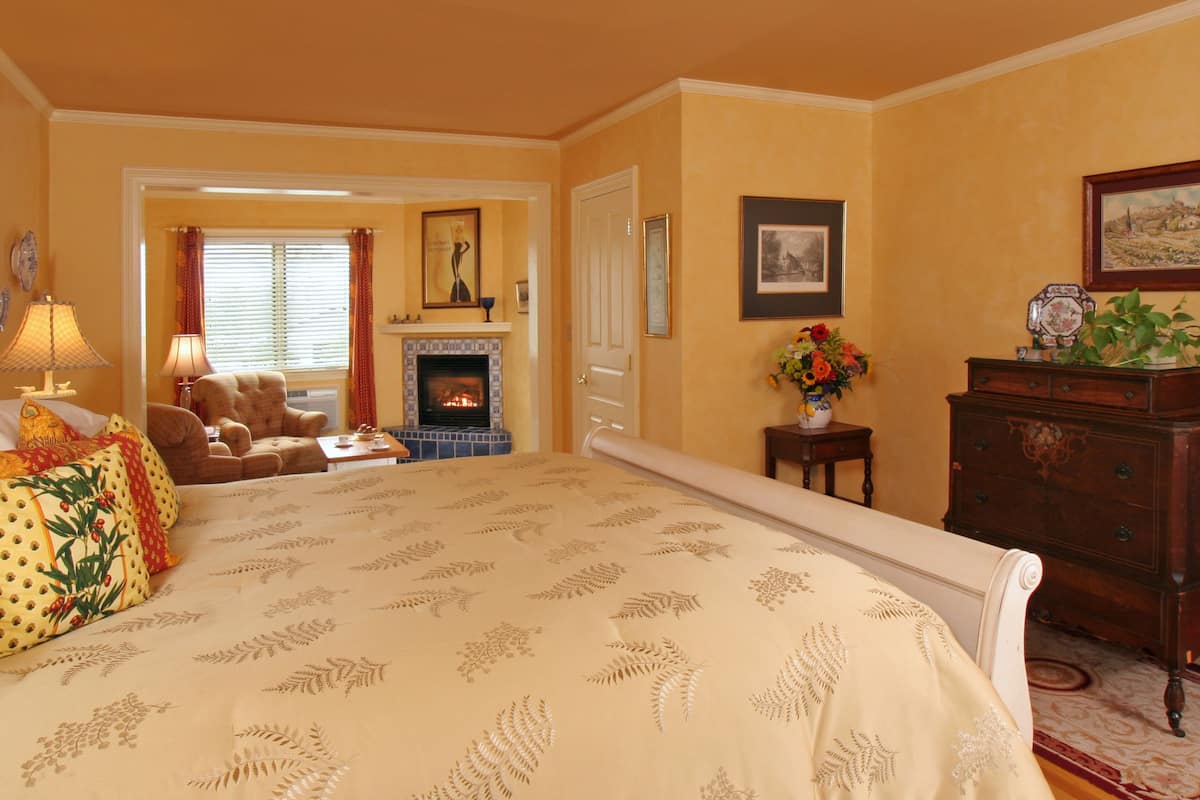 A spacious bedroom with a large bed, a fireplace, a sitting area with a chair and table, and a dresser with a vase of flowers.