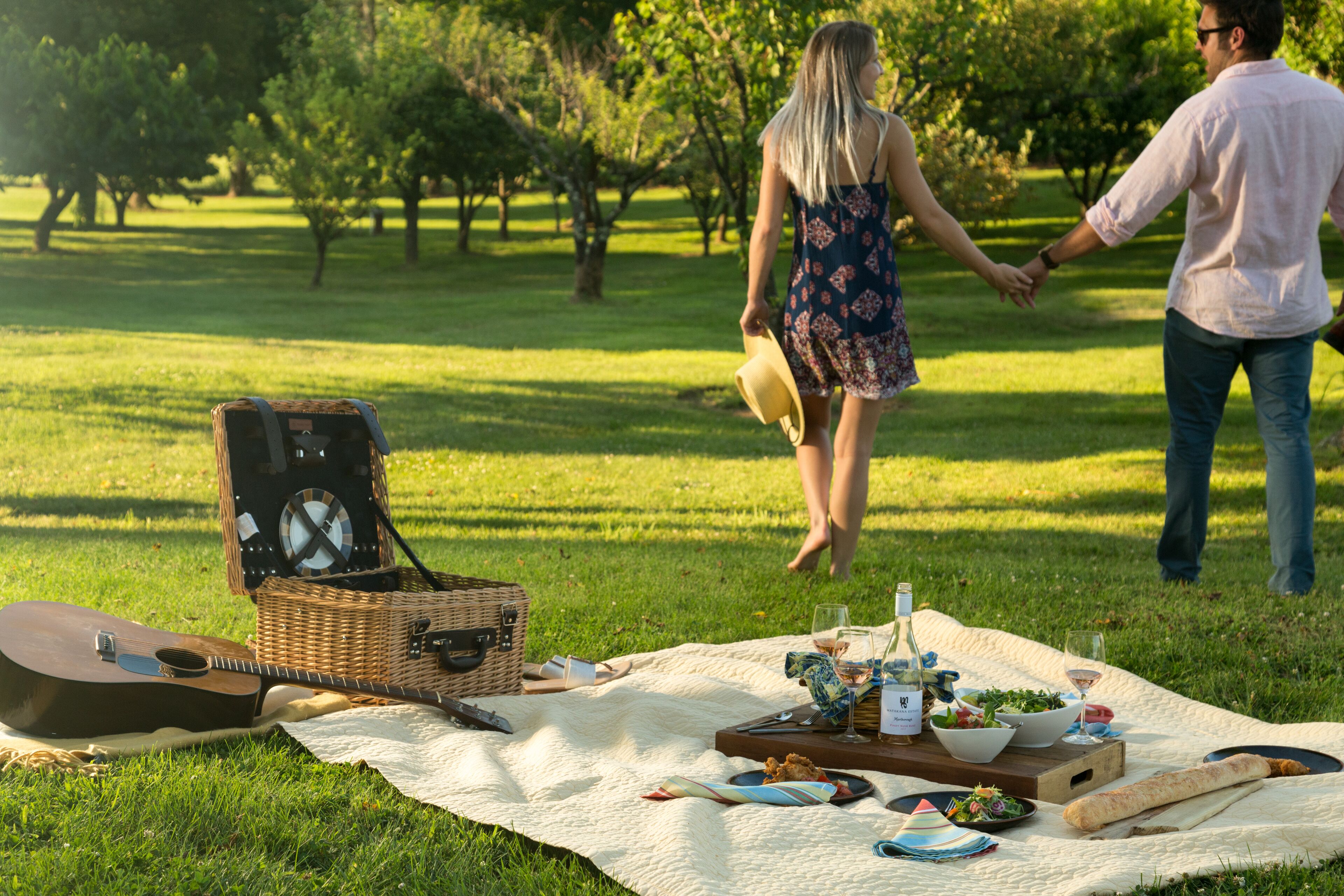 A picnic setup with a guitar, picnic basket, and food on a blanket in a grassy area.