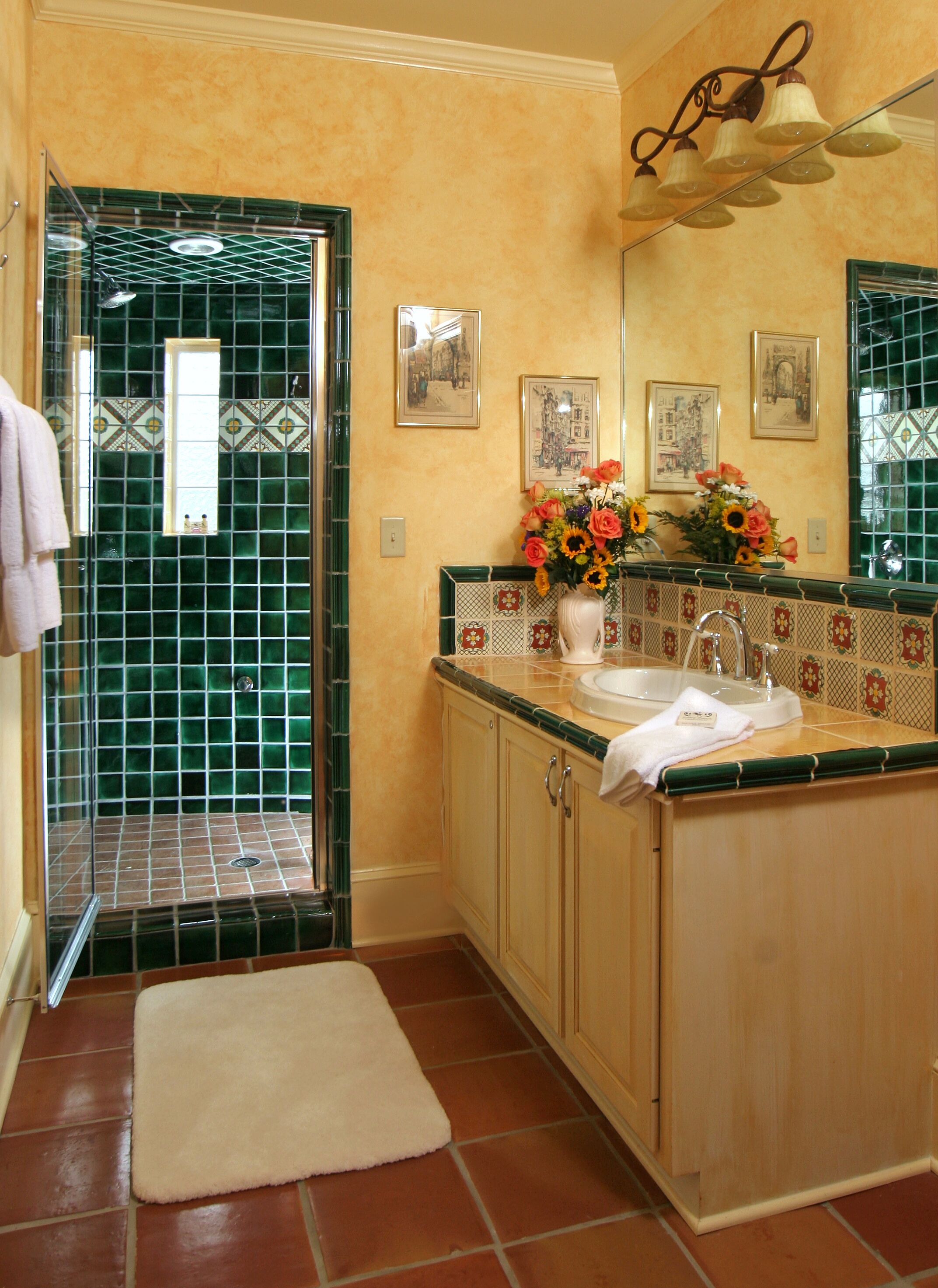 A bathroom with a green-tiled shower, a sink with a mirror above it, and a vase of flowers on the counter.