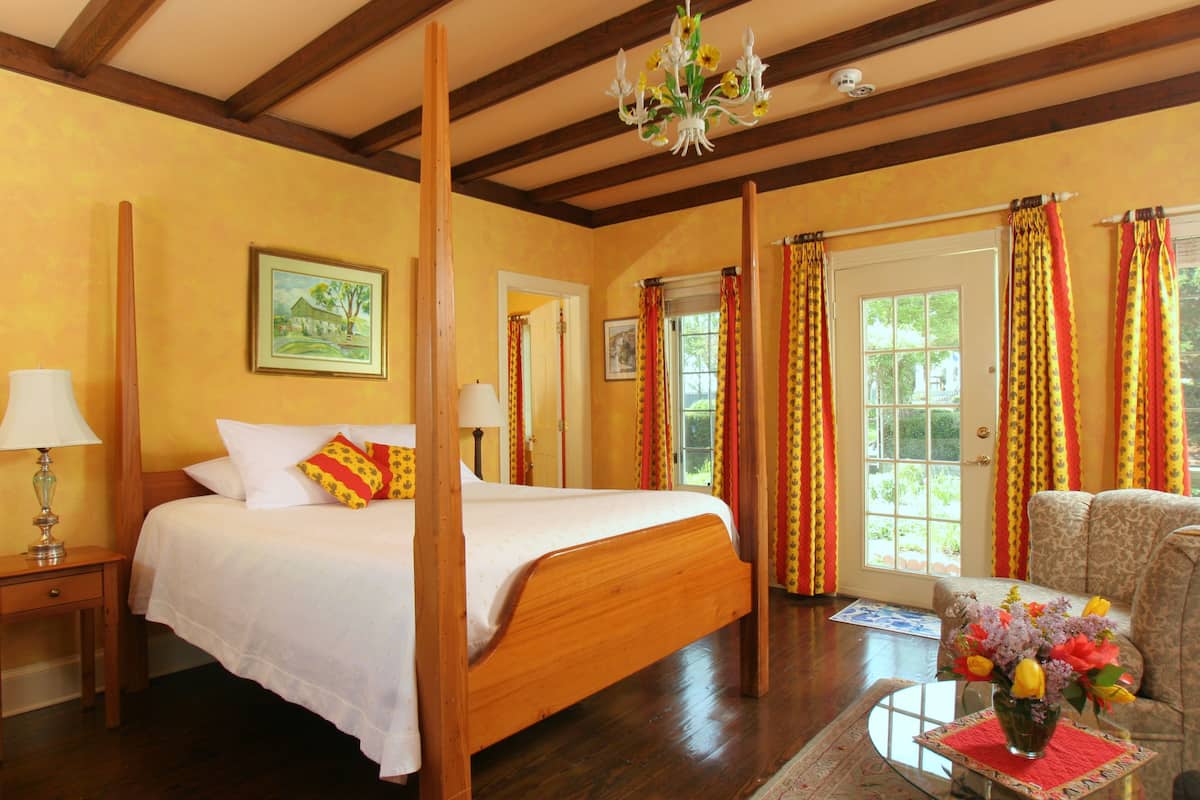 A four-poster bed with white linens, a wooden nightstand with a lamp, a floral vase on a table, and a patterned armchair.