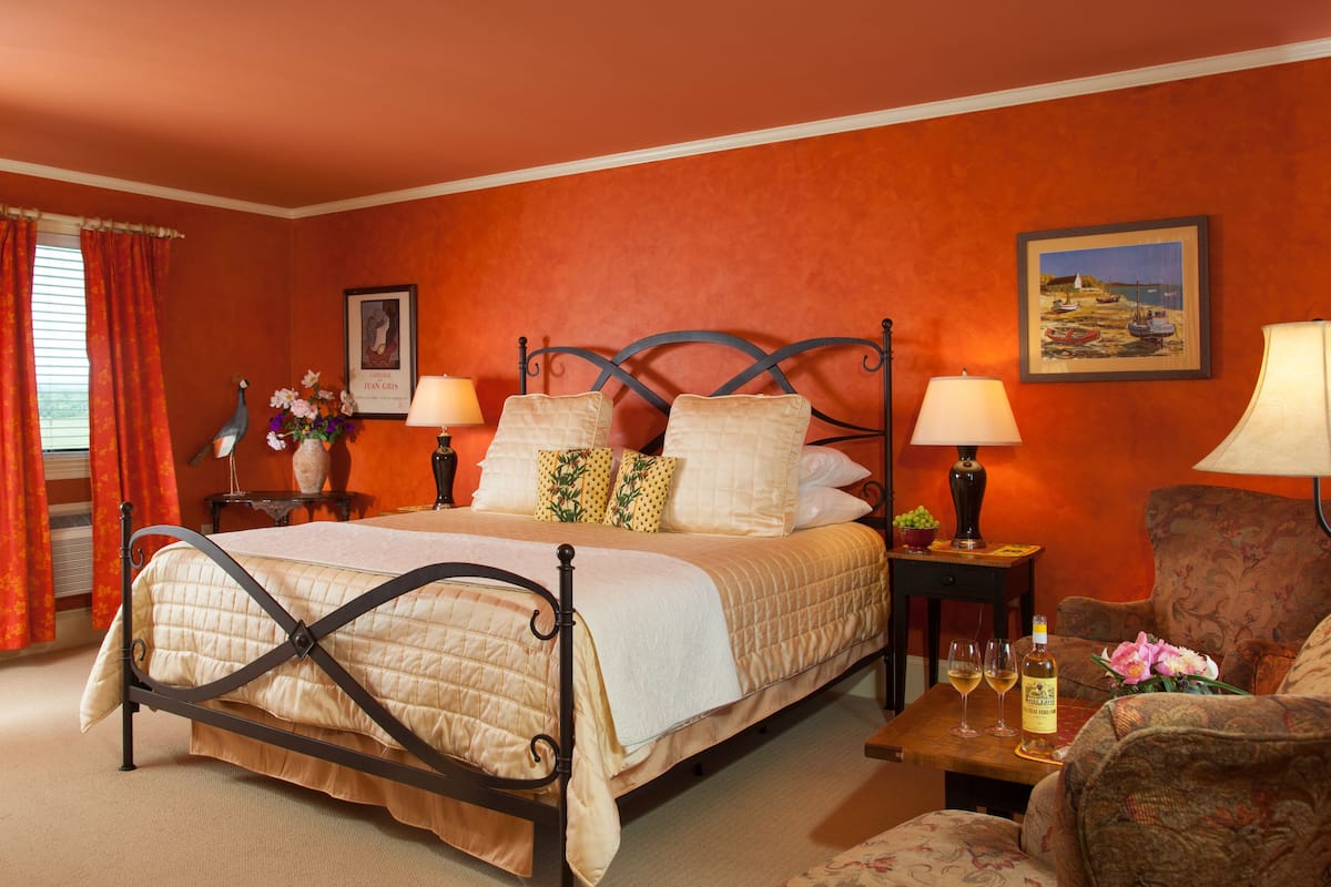A hotel room with a bed, two bedside tables, a chair, a window with curtains, a framed picture on the wall, and a small table with a bottle and glasses.