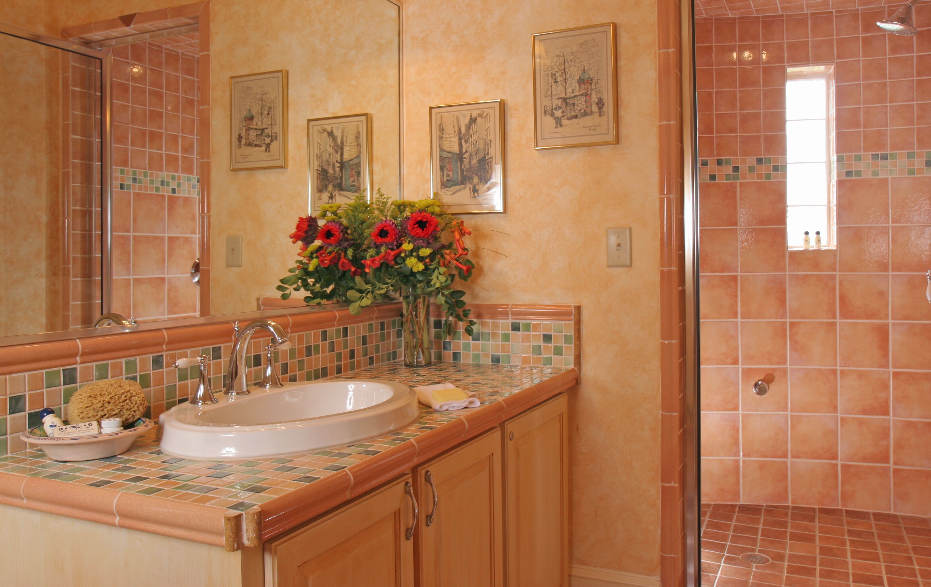 A bathroom with a large mirror, a sink, and a shower. There are framed pictures on the wall and a vase of flowers on the counter.