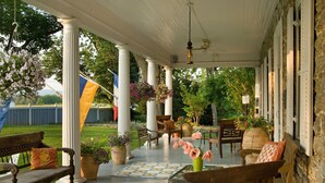 A covered porch with wooden benches and potted plants, a tiled floor, and a view of a grassy yard. - L'Auberge Provencale Inn & Restaurant (Boyce)